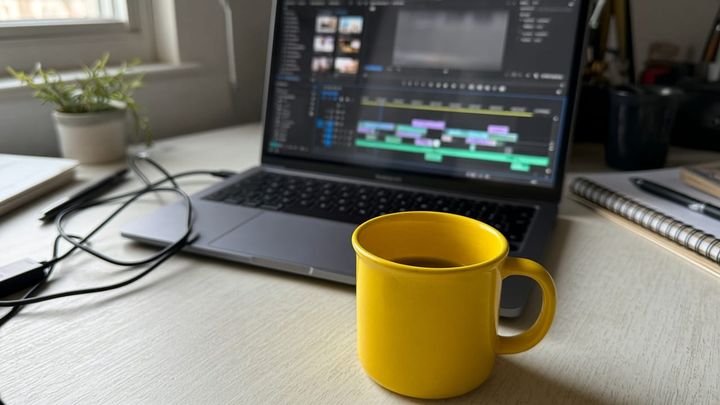 A laptop showing a blurred video-editing timeline on a plain desk beside a sunflower-yellow enamel mug, handheld iPhone photo in late afternoon window light.