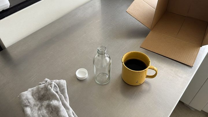 A new unlabeled screw cap and a plain glass bottle sitting together on a scratched stainless steel prep bench in flat window light, beside a dented golden yellow enamel mug of black coffee, 
