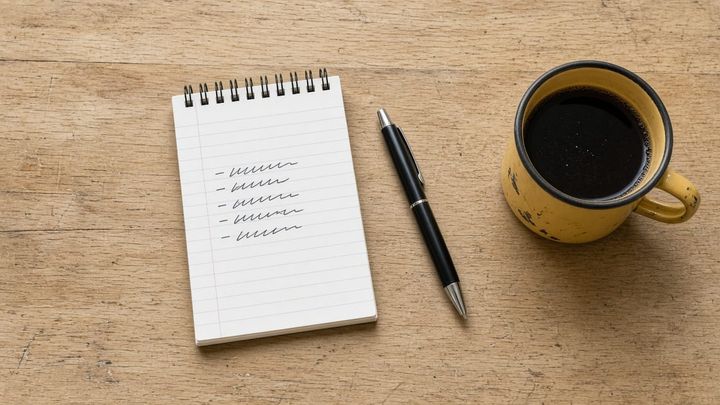 Overhead flat lay of a plain lined notepad on a scratched pale butcher block counter in flat morning window light, a ballpoint pen laid beside it and a dented golden yellow enamel mug of bla