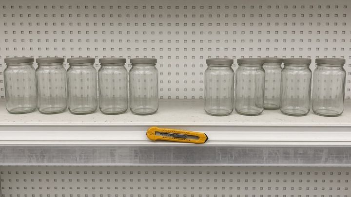 Eye level phone photograph of one section of a plain grocery store shelf, a row of unlabeled glass jars with an obvious empty gap where several have already been taken, and a dented golden y