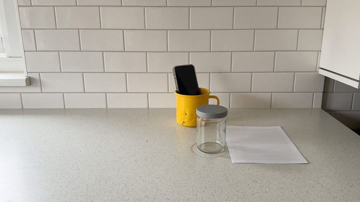 Eye level phone photograph of a kitchen counter with a phone propped on a golden yellow enamel mug filming a single open jar, a paper invoice lying flat beside it, crumbs on the counter, ord