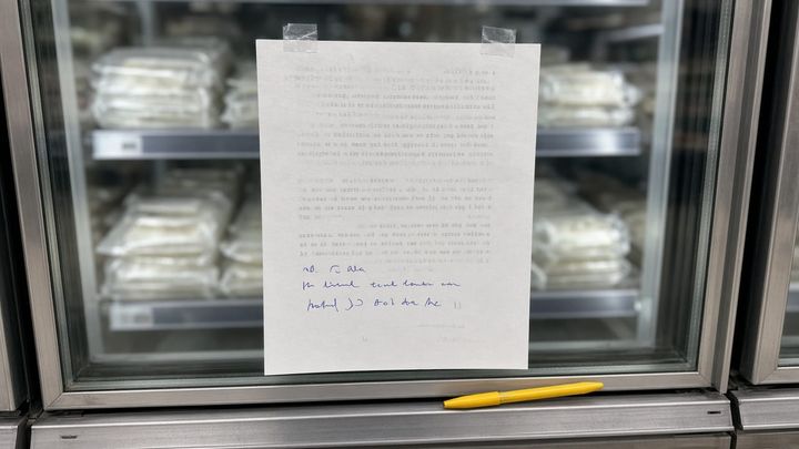 Eye level phone photograph of a plain sheet of paper taped to a refrigerated grocery case, a golden yellow ballpoint pen resting on the case ledge beside it, ordinary fluorescent store light