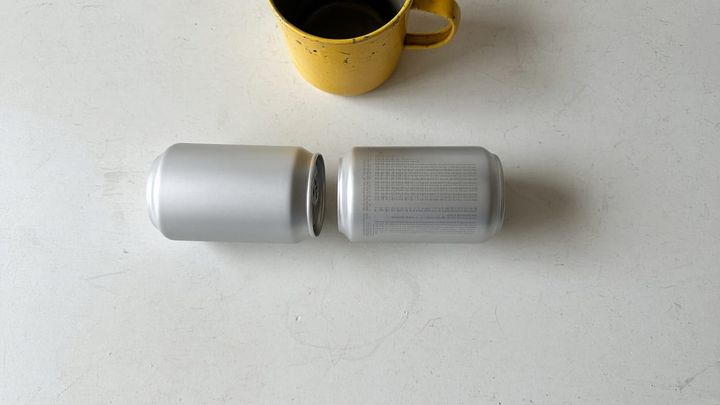 Overhead photograph of two plain unlabeled silver beverage cans on a scuffed white counter, one turned to its blank front and one turned to a block of illegible fine print, with a dented gol
