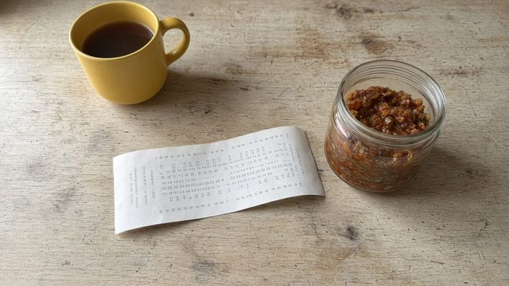 An overhead phone photo of a curled paper grocery receipt on a wood kitchen counter next to a plain glass jar and a yellow coffee mug in morning light, no logos.