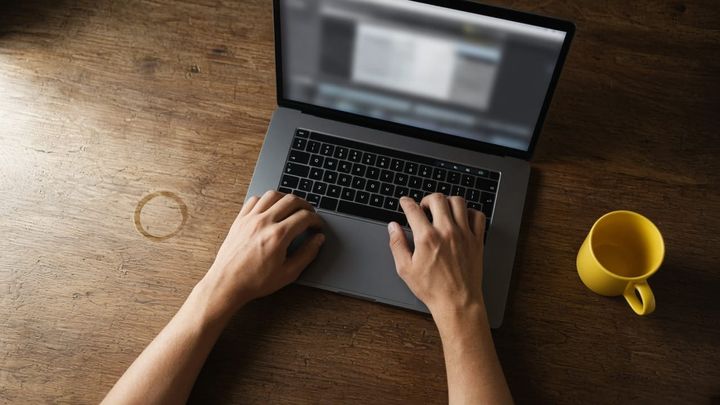 Overhead shot of hands pausing a video edit on a laptop trackpad at a worn wood desk, a yellow mug beside it, morning window light, no logos, no readable screen text.