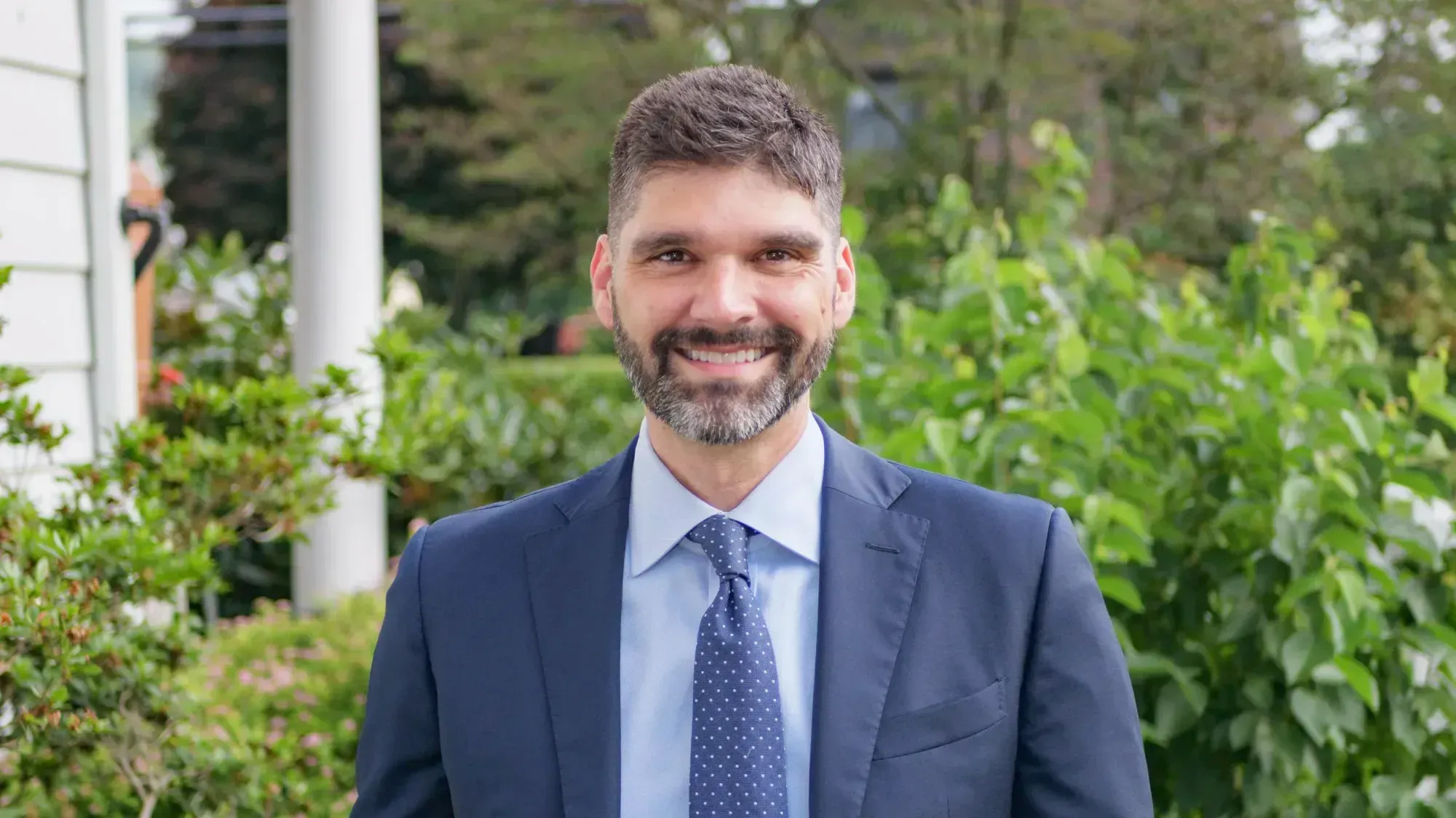 A head and shoulders photo of a smiling Aaron Gies in blue business suit. He is standing outside with bushes in the background.