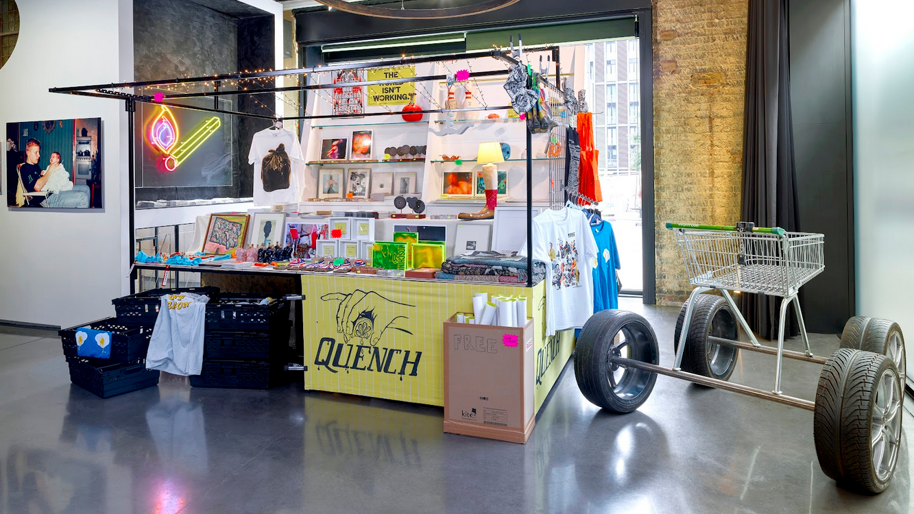 Merch kiosk with bin-bag tees and ceramic notes beside Grace Clifford’s bronze Big Shop.