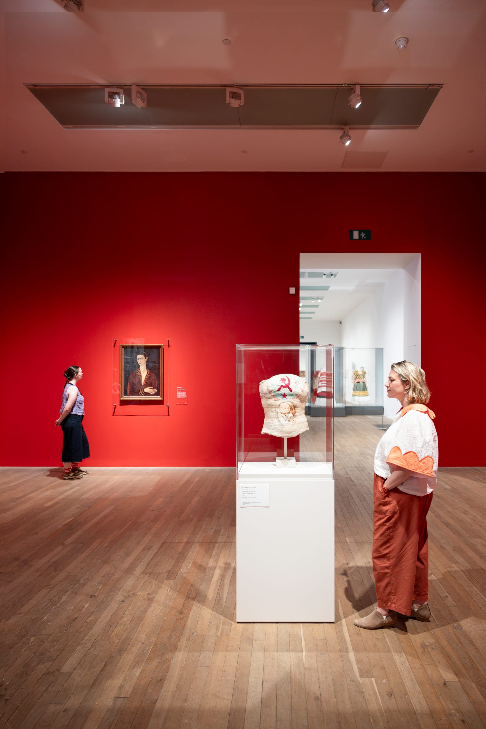 A red gallery wall with a Frida Kahlo painting and a glass case containing a painted medical corset, viewed by visitors.