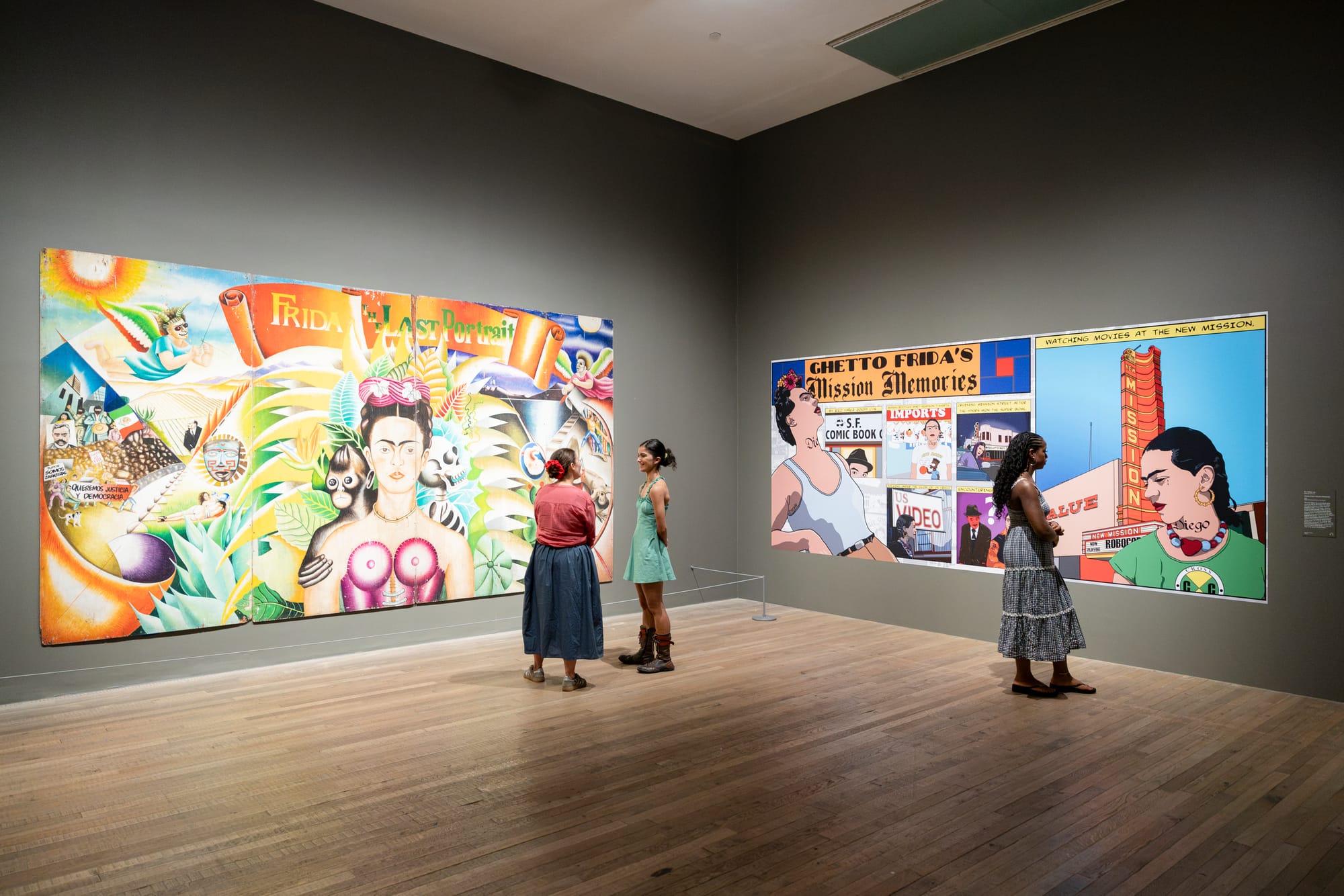 Visitors view large colourful artworks in a grey-walled gallery, including later artistic responses to Frida Kahlo’s image.