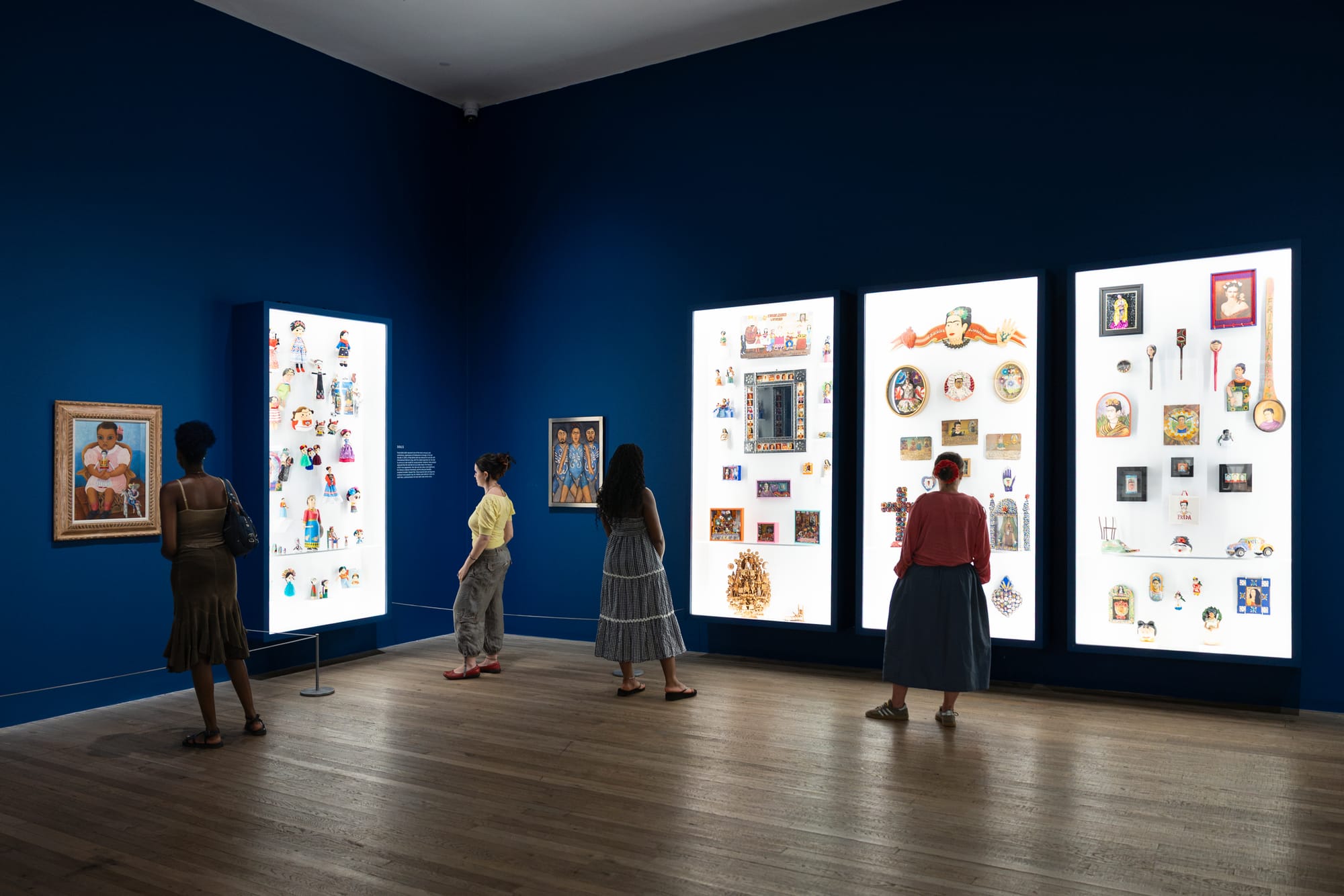 Visitors stand in a dark blue gallery facing illuminated display cases filled with small objects and images related to Frida Kahlo.