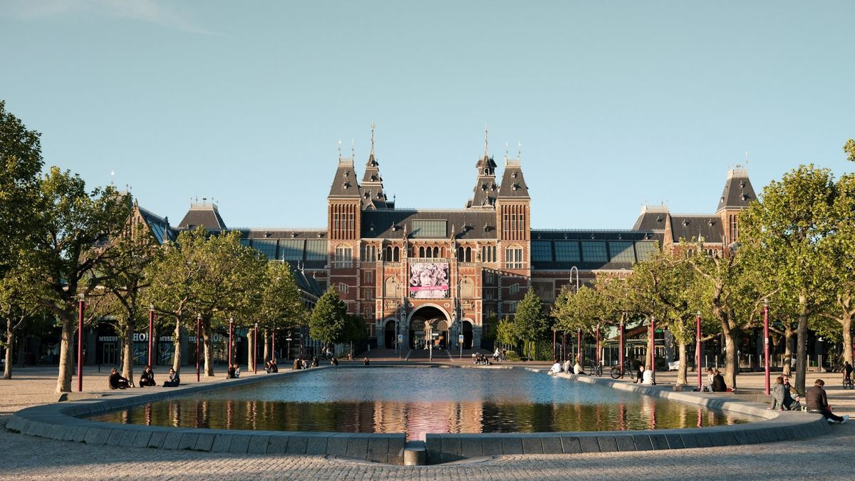 Exterior of the Rijksmuseum in Amsterdam, with its brick arches and twin towers rising above cyclists and pedestrians moving across Museumplein.