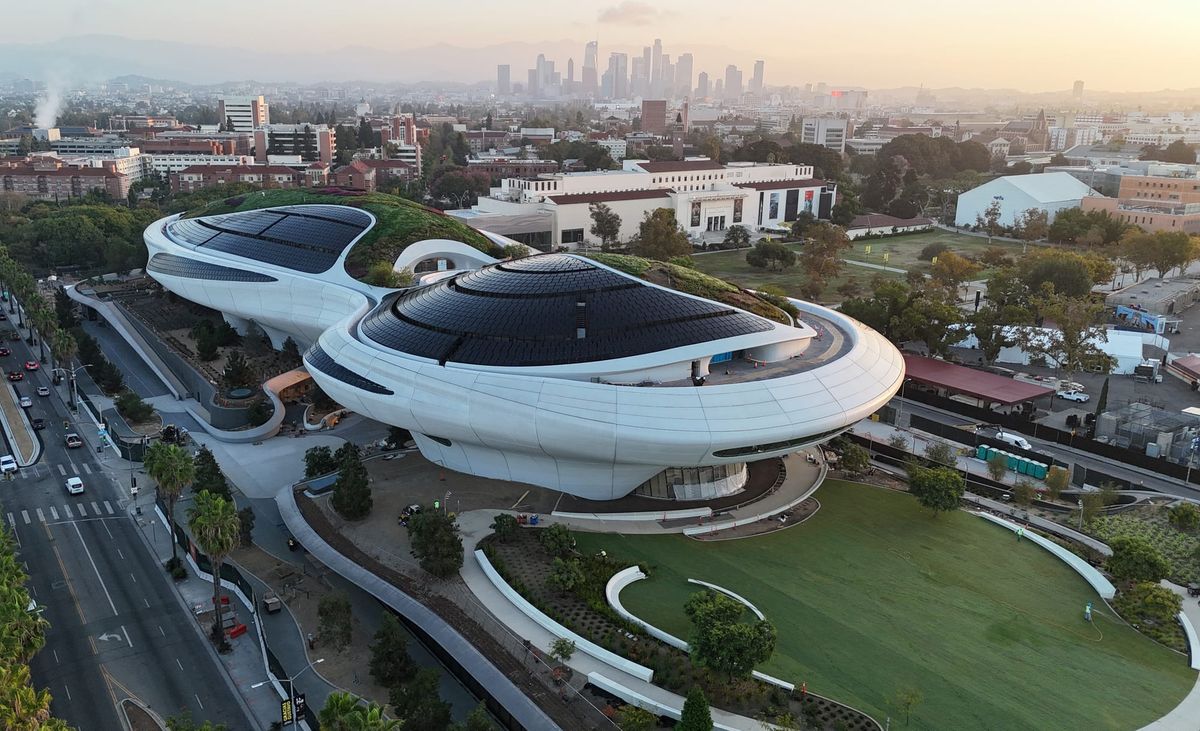 Aerial view of the under-construction Lucas Museum of Narrative Art in Exposition Park, a curved white building surrounded by cranes with the Los Angeles skyline in the background.