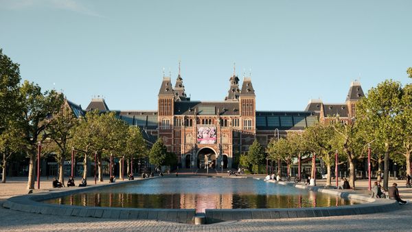 Exterior of the Rijksmuseum in Amsterdam, with its brick arches and twin towers rising above cyclists and pedestrians moving across Museumplein.
