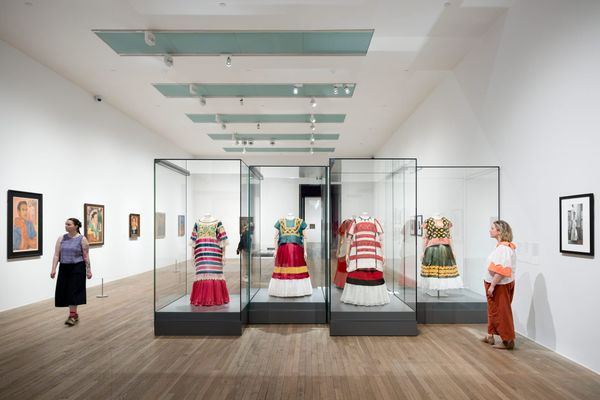 Visitors in a white-walled gallery view Frida Kahlo paintings and glass cases displaying colourful garments from the exhibition at Tate Modern.