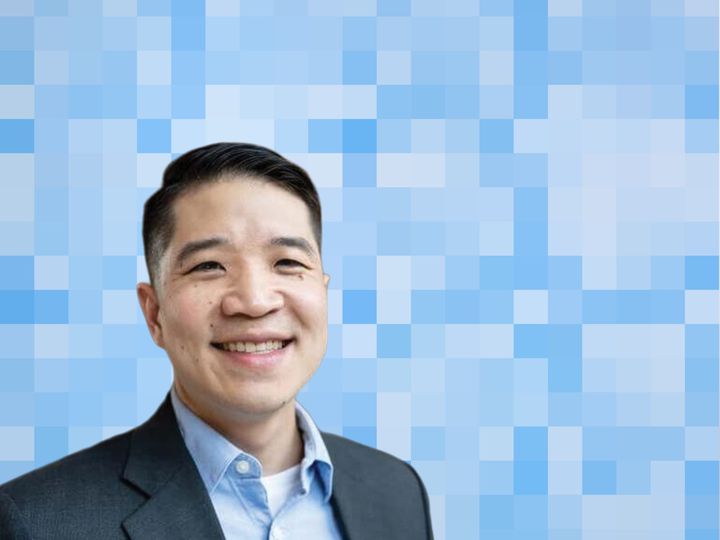 Jeremy Lee, wearing a dark suit jacket over a blue collared shirt, smiles at the camera in front of a stylized blue pixel-grid backdrop.
