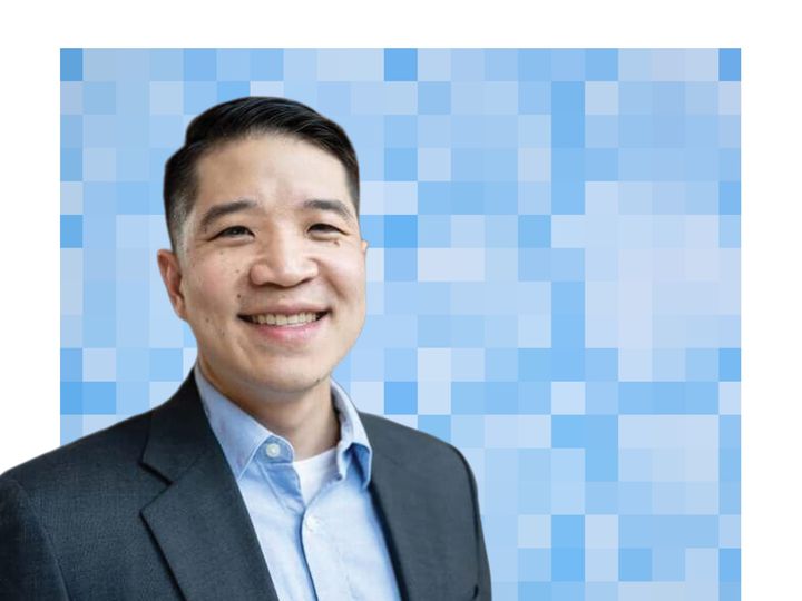 Jeremy Lee, wearing a dark suit jacket over a blue collared shirt, smiles at the camera in front of a stylized blue pixel-grid backdrop.