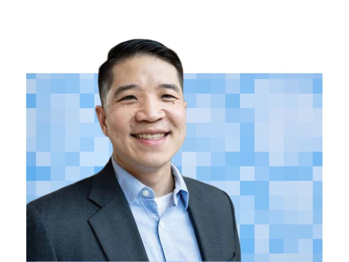 Jeremy Lee, wearing a dark suit jacket over a blue collared shirt, smiles at the camera in front of a stylized blue pixel-grid backdrop.