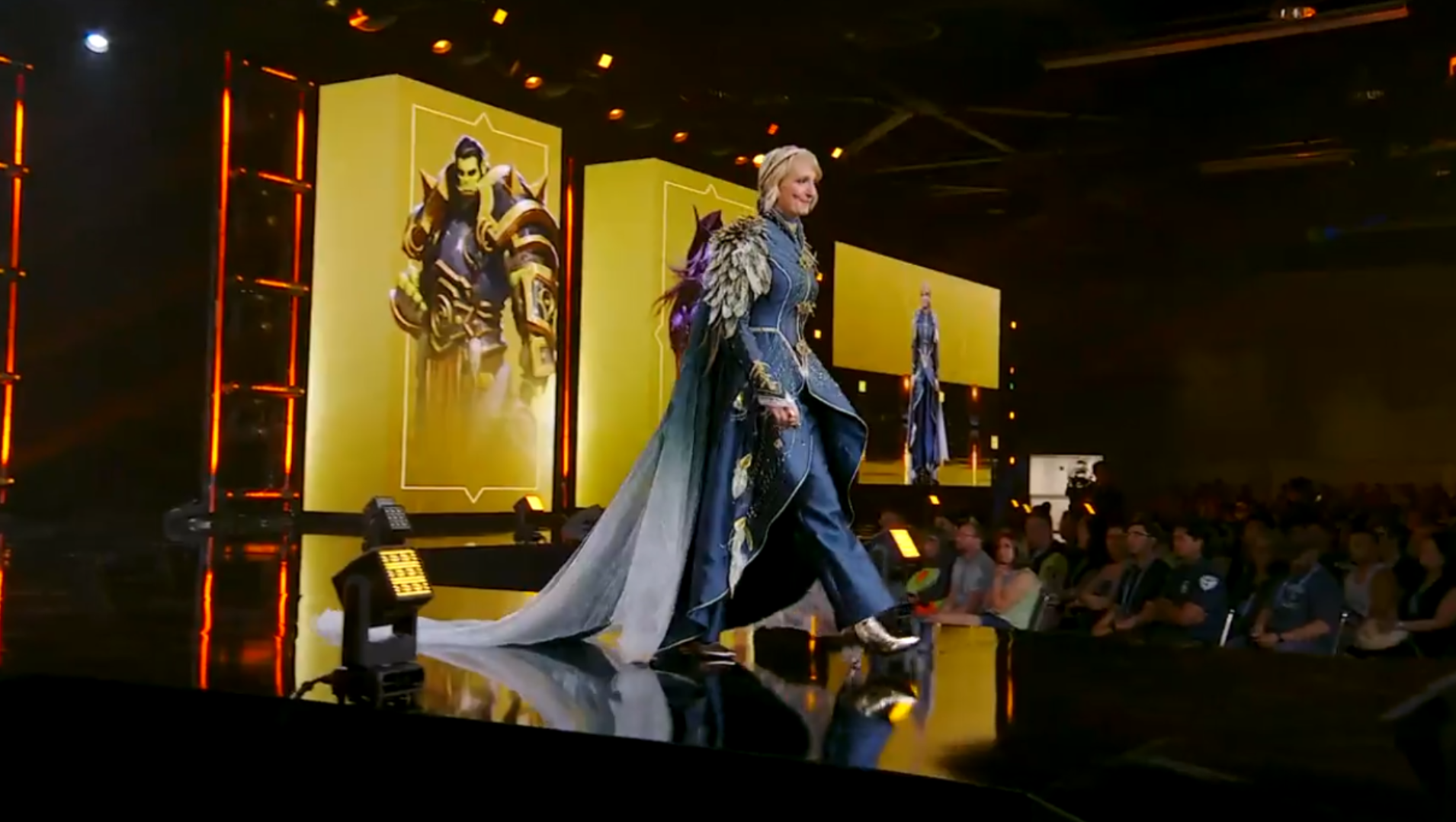 Holly Longdale wearing an elaborate blue and gold coat, with feathered pauldrons and a long silvery cape.