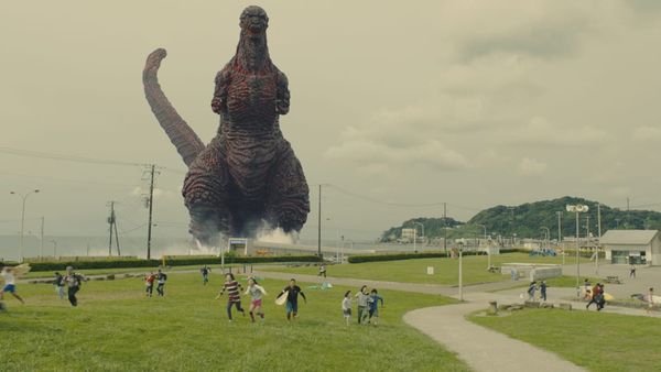 An image from Shin Godzilla (2016) with Godzilla walking ashore in the background while people run in the foreground.