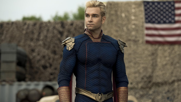 Homelander, the antagonist of The Boys, stsands in front of a military background with an American flag. He's a handsome white man with blond hair, a blue super suit, and golden eagle orname