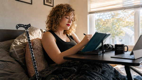 The Chronic Worker stock photo og disabled woman working from bed with a skull cane and teal journal.