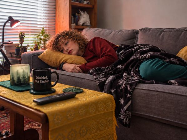 A woman rests on a gray couch under a patterned blanket, with a mug reading “Disabled, Not Unable” on the coffee table.