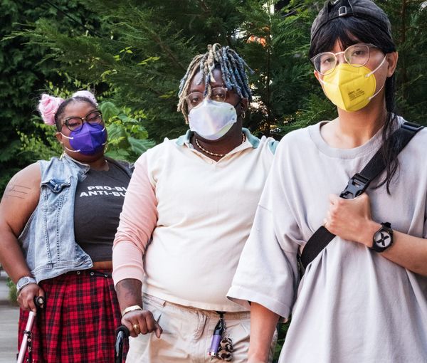 Three disabled people of color wearing face masks stand together outdoors with two canes and a forearm crutch against dark green foliage.