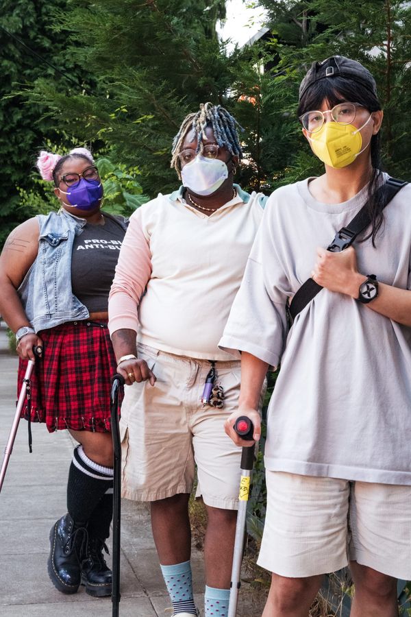 Three disabled people of color wearing face masks stand together outdoors with two canes and a forearm crutch against dark green foliage.