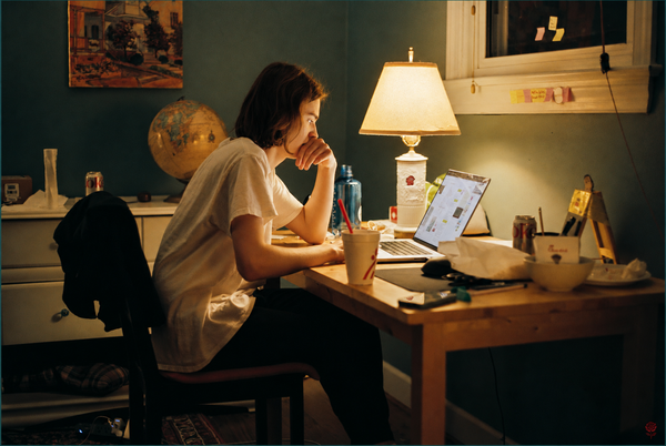 A woman sits at a cluttered desk in a dim room, leaning toward her laptop with one hand near her mouth as a lamp glows beside her.