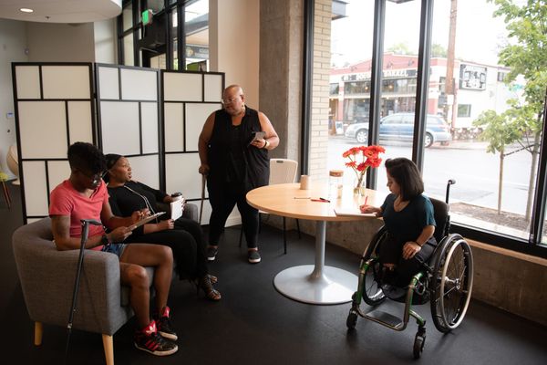Four disabled people of color gather in an open circle for a meeting; two people use canes, one person uses a wheelchair and takes notes, and another stands with a tablet.