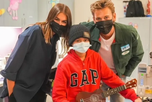 Kaia Gerber and Austin Butler wearing face masks in hospital, pose with a young patient wearing a red jumper and a facemask t