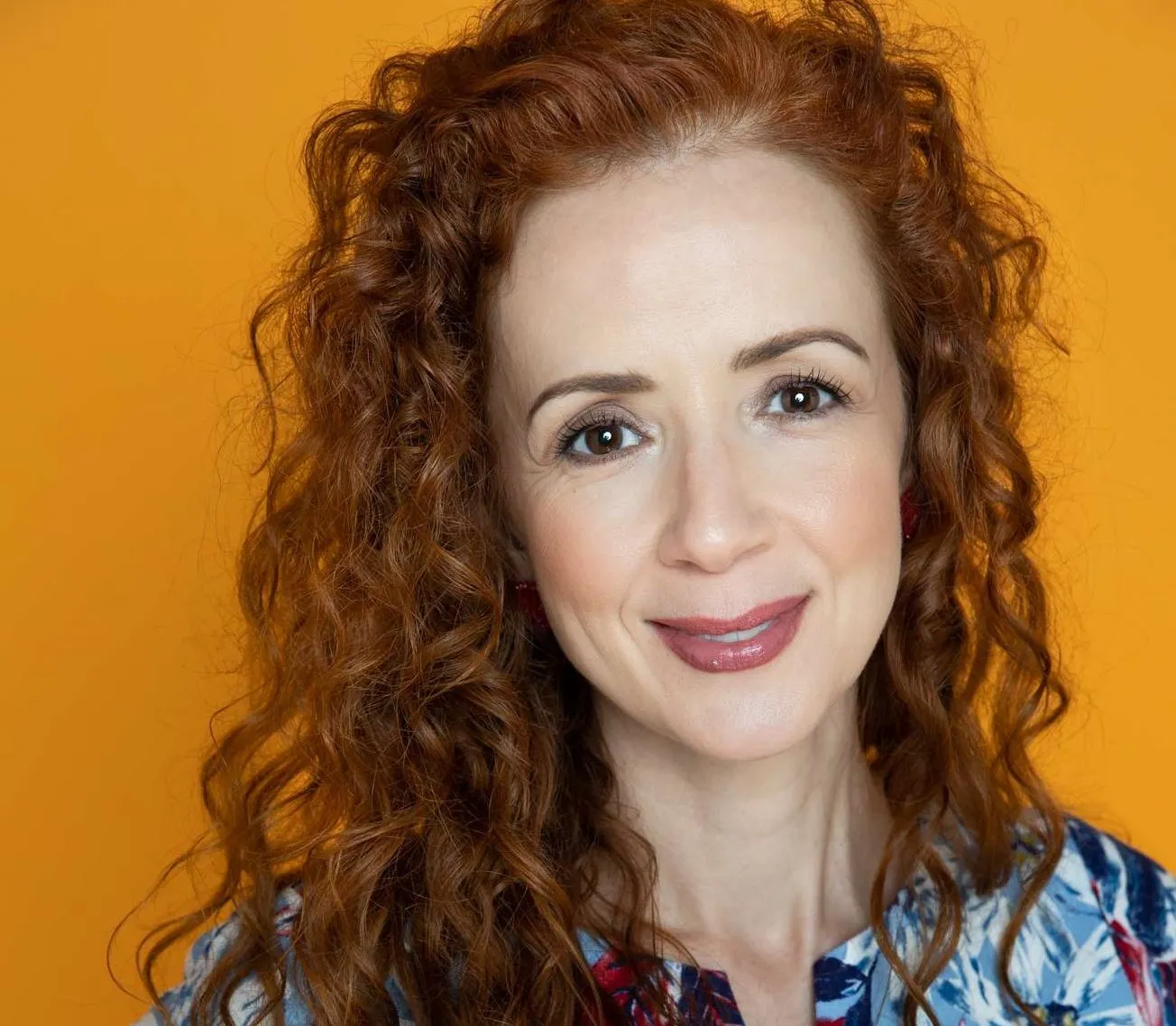 Ana Maria Belo smiling. She has curly red hair and stands in front of an orange background.