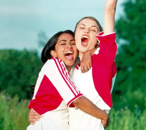 Parminder Nagra and Keira Knightley as Jess and Jules in white and red uniforms celebrating on the field in Bend It Like Beck