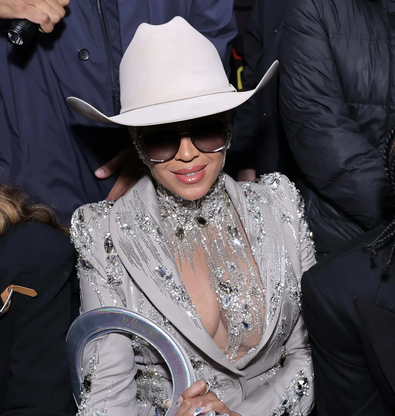 Beyoncé wears a cowboy hat and sunglasses as well as a silver rhinestone jacket. She's smiling at a red carpet event.