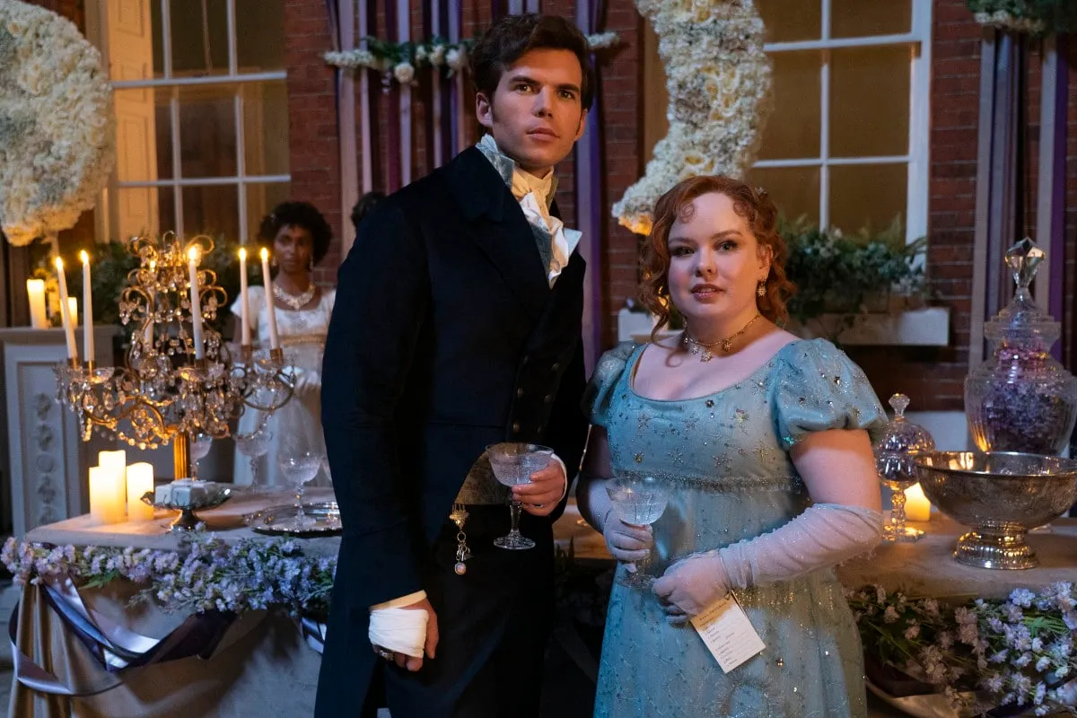 a still of Colin and Penelope in Bridgerton season 3. They are looking surprised but composed and are surrounded by wealthy t