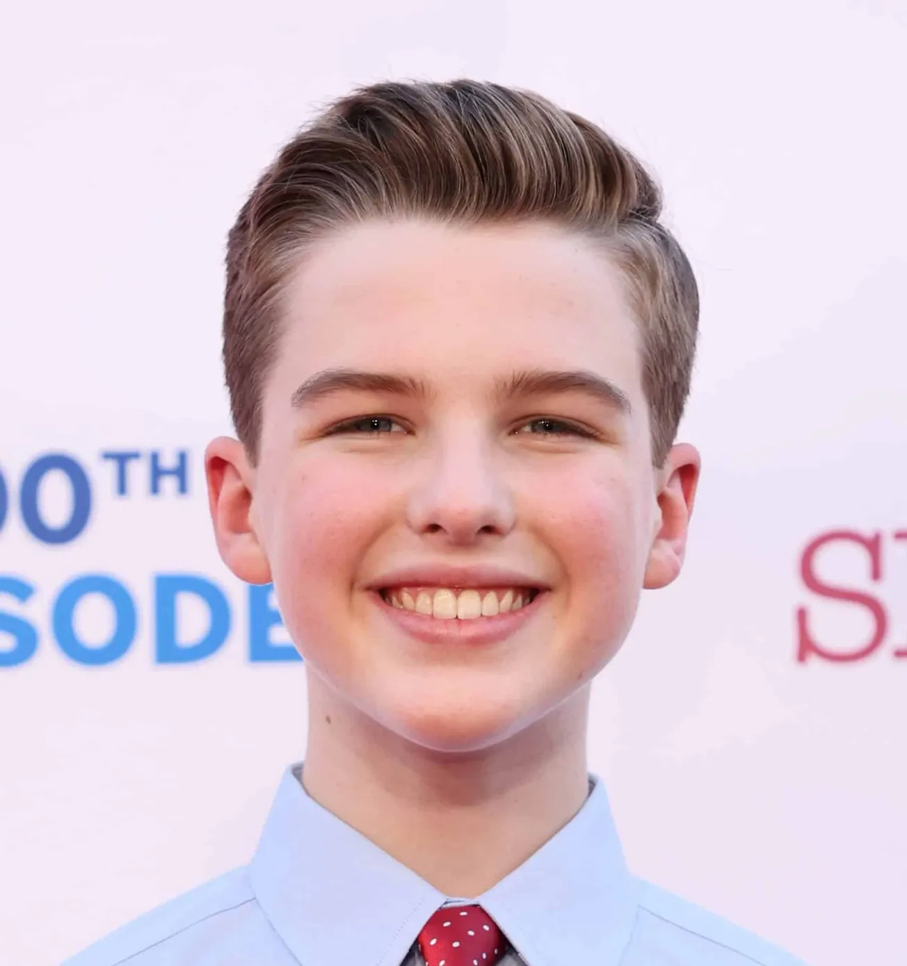Iain Armitage, star of 'Young Sheldon' smiles wearing a blue shirt and red tie at a press event in 2022.
