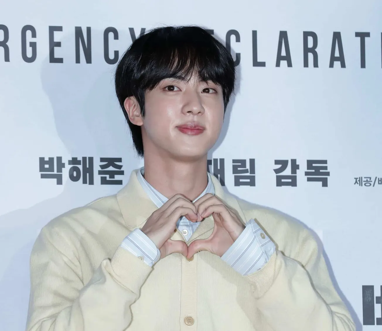 Jin of BTS makes a heart shape with his hands at a press event. He wears a cream jumper and stands in front of a white wall w