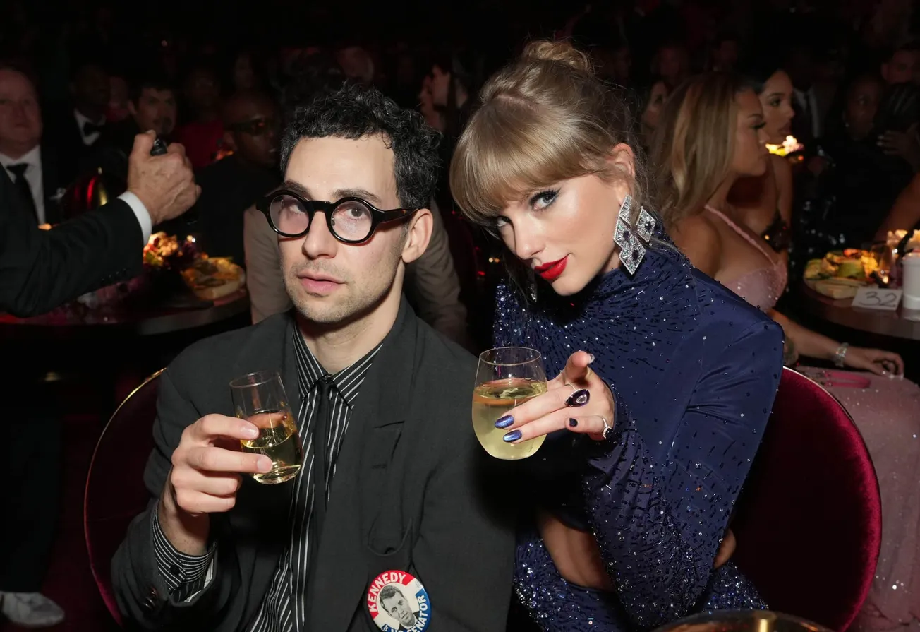 Jack Antonoff and Taylor Swift holding drinks and sitting at a table, looking directly at the camera.