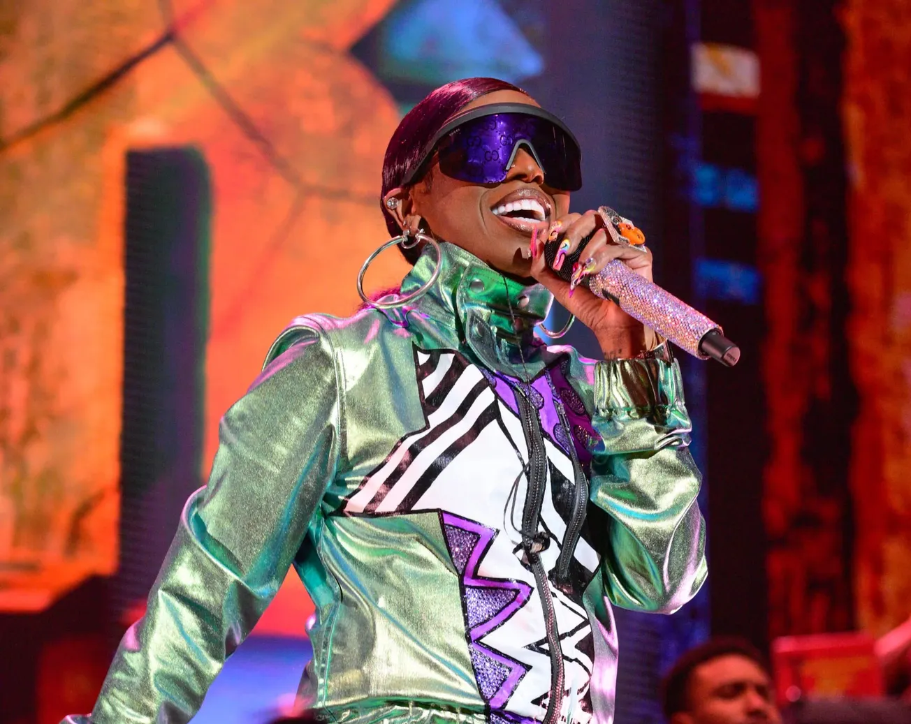 Missy Elliot wears shiny greet jacket and large sunglasses holding a mic to her mouth as she performs on stage.