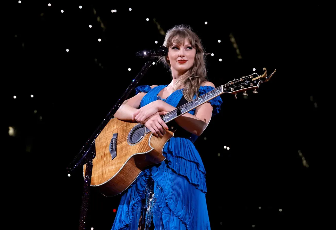 Taylor Swift holds her guitar on stage and wears a blue dress during her Eras tour concert in Los Angeles.