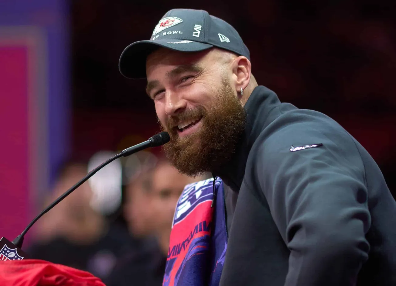 a close up photo of Travis Kelce sitting at a press conference and smiling while he speaks into the mic. He's wearing a Super