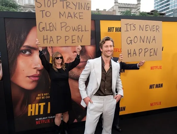 Glen Powell attends the premiere of Hit Man wearing a cream suit and brown shirt. His parents stand behind him holding funny