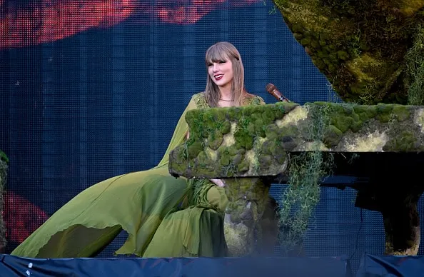 Taylor Swift dressed in a flowing green dress and sitting at her piano on stage which is covered in greenery.