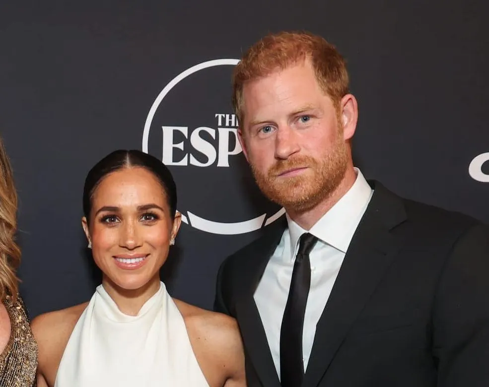 Harry and Meghan smiling at a press event. SHe wears a halter cream dress and he wears a suit. Today they've launched The Par