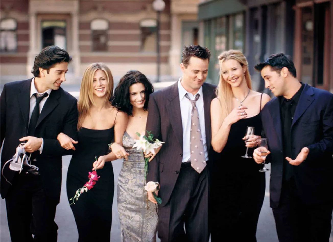Friends cast photo walking down a street, arm in arm, smiling.