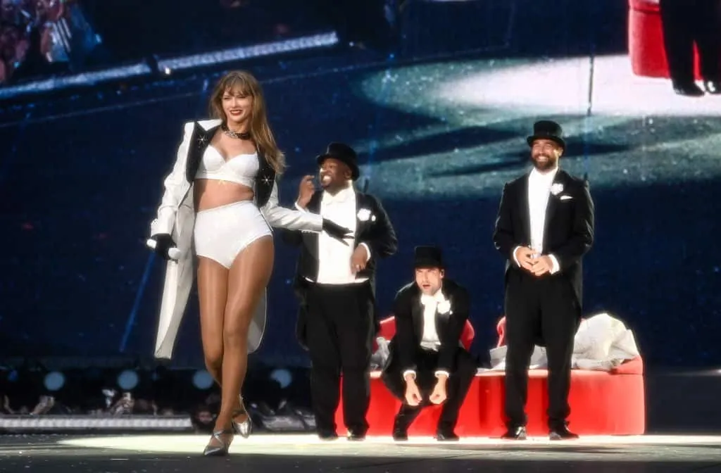 Taylor Swift on stage smiling with her backup dancers dressed in top hats and tails behind her. One of them is Travis Kelce!