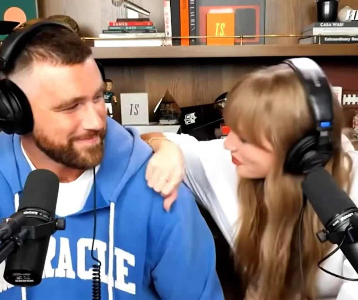 A still of Travis Kelce and Taylor Swift during the New Heights podcast, They're looking at each other, smiling and Taylor ha