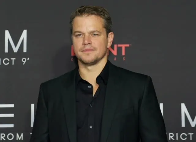 Matt Damon smiles on the red carpet dressed in a black shirt and suit jacket.