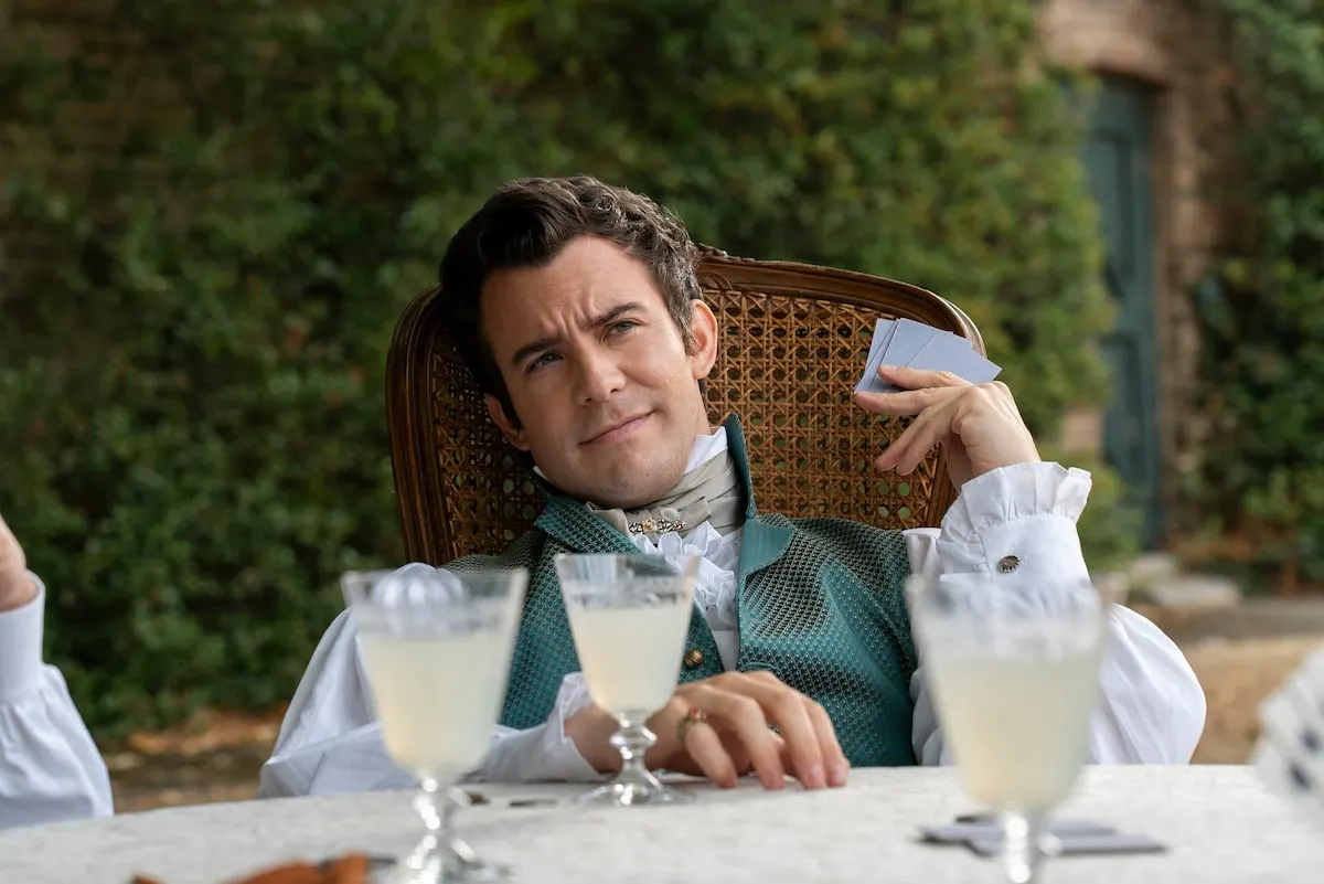 Luke Thompson as Benedict Bridgerton sitting at a table holding up some cards.