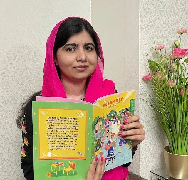 Malala wears a pink scarf and holds up a copy of her "assembly" magazine.